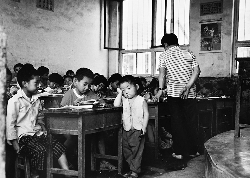 1995年，济南南部山区一所民办小学里的一位老师正在上课，旁边的这位孩子是这位老师的孩子。或许因为老师家庭的原因，老师为了不耽误给学生上课无奈把自己的孩子带到了课堂上。作者：李瑞勇